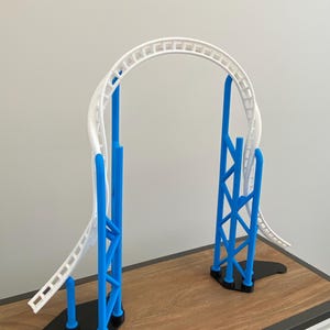 Könnte beinhalten: Ein Miniatur-Achterbahnmodell mit weißer Strecke und blauen Stützstrukturen. Die Strecke bildet eine Schleife, die von blauen vertikalen Trägern und schwarzen Basen getragen wird. Das Modell wird auf einer Holzoberfläche präsentiert.