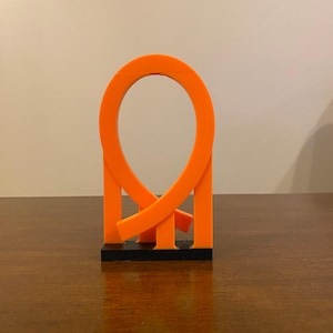 Puede incluir: Una escultura abstracta naranja con un diseño de bucle, montada sobre una base rectangular negra. La escultura tiene una estética moderna y minimalista, adecuada para la decoración del hogar o la oficina. El color naranja brillante proporciona un contraste visual llamativo.