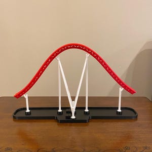 Pode incluir: Um modelo em miniatura de montanha-russa com uma pista vermelha e estruturas de suporte brancas. A pista curva-se em arco, sustentada por vigas brancas verticais e anguladas. A base é preta, sobre uma superfície de madeira.