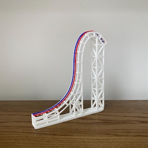 Puede incluir: Modelo blanco impreso en 3D de una montaña rusa con pistas rojas y azules. La montaña rusa presenta un bucle y una estructura de soporte con un diseño de celosía. El modelo se muestra sobre una superficie de madera.