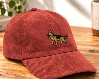 May include: A rust-colored corduroy baseball cap featuring an embroidered graphic of a German Shepherd dog. The dog is depicted in profile with tan and black fur, standing on all fours. The cap has a structured crown and a curved brim.