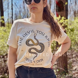 May include: Cream-colored t-shirt with the text "THE DEVIL YOU KNOW" encircling a snake illustration. Below, the text "DEVIL'S DEN Gettysburg, Penna." is printed. The shirt is worn with dark jeans and sunglasses.