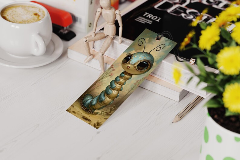 May include: A whimsical bookmark featuring a cartoon caterpillar with large, expressive eyes. The caterpillar is primarily blue and gold, set against a soft, illustrated background. The bookmark is rectangular with a small hole at the top.