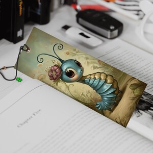 May include: A whimsical bookmark featuring a cartoon caterpillar with large eyes, blue segmented body, and a flower. The bookmark is inserted into a book, with a black and gold cord attached.