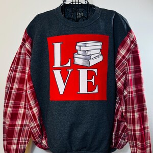 May include: A gray sweatshirt with red and white plaid sleeves. The front of the sweatshirt has a red square with the word "LOVE" in white letters and a stack of books in the middle of the word.