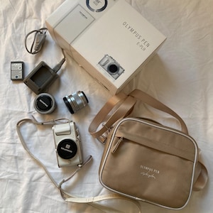 Könnte beinhalten: Ein Olympus PEN E-PL9 Kamera-Kit, einschließlich einer weißen Kamera mit Objektiv, einer beigefarbenen Kameratasche, einer Box und Zubehör. Die Kameratasche hat einen Schultergurt und die Aufschrift "Olympus PEN".