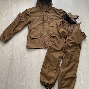 Könnte beinhalten: Ein braunes Outdoor-Jacken- und Hosen-Set. Die Jacke hat eine Kapuze, mehrere Taschen und Knopfverschlüsse. Die Hose hat einen Kordelzug an den Knöcheln. Das Set enthält ein Paar schwarze Handschuhe. Das Outfit besteht aus strapazierfähigem Material.