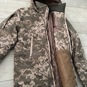 May include: A hooded camouflage jacket with a zip and pockets. The jacket has a pixelated design in shades of green, brown, and beige. The interior lining is a solid light brown colour. The jacket is designed for outdoor wear and protection from the elements.
