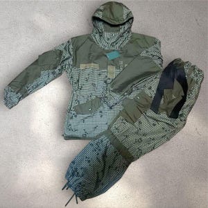 Può includere: Un completo mimetico a due pezzi nei toni del verde e del bianco. La giacca ha un cappuccio, più tasche e un motivo a scacchi. Anche i pantaloni presentano tasche e un design a scacchi abbinato. Il completo è progettato per l'uso all'aperto.
