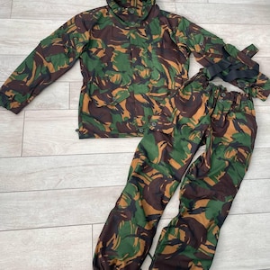 Peut inclure: Ensemble veste et pantalon de camouflage vert, marron et beige. La veste a une capuche et des boutons. Le pantalon a une taille élastique et des boutons. L'ensemble est fait d'un tissu résistant.