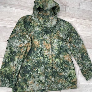 Op de afbeelding: Een camouflagejas met capuchon, met een patroon van groene, bruine en grijze vlekken. De jas heeft meerdere zakken en een knoopsluiting aan de voorkant. De stof lijkt een duurzaam, outdoor-klaar materiaal te zijn.