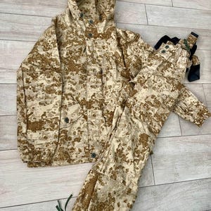 Puede incluir: Conjunto de chaqueta y pantalón de camuflaje beige y marrón. La chaqueta tiene capucha y botones. El pantalón tiene bolsillos y cordones. El conjunto está hecho de un material resistente y está diseñado para uso en exteriores.