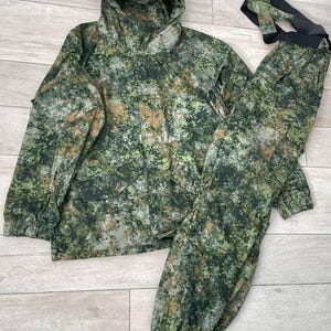May include: A camouflage hunting suit in shades of green, brown, and gray. The set includes a hooded jacket with pockets and matching pants with suspenders. The fabric has a pixelated pattern.