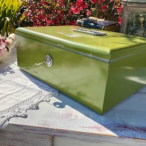 Puede incluir: Una caja de metal verde vintage con una cerradura y un pestillo plateado. La caja está sobre una superficie blanca con un mantel de encaje.