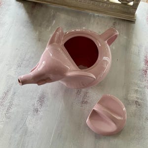 May include: A pink ceramic teapot shaped like an elephant with a separate lid. The teapot has a spout and a handle. The lid has a small handle.