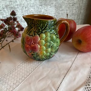 Könnte beinhalten: Eine Keramikkanne mit braunem Rand und einem grünen, roten und weißen Design aus Trauben und Kirschen. Die Kanne steht auf einer weißen Tischdecke mit Spitzenbesatz.