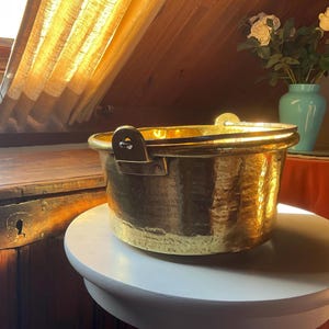 May include: A large, antique brass pot with two handles. The pot has a hammered finish and is sitting on a white surface.