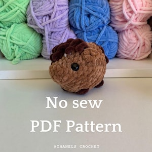 Puede incluir: Un conejillo de indias de amigurumi tejido a crochet marrón con ojos negros. El conejillo de indias está sentado sobre una superficie blanca con ovillos de lana en el fondo. El texto "No sew PDF Pattern" está escrito debajo del conejillo de indias. El texto "@CHANELS CROCHET" está escrito debajo del texto.