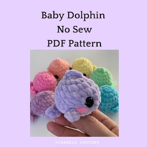 May include: A close-up of a purple crocheted baby dolphin toy. The toy is being held in a hand. The toy is part of a set of colorful crocheted baby dolphin toys. The image includes the text "@CHANELS_CROCHET".