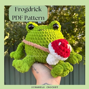 May include: A green crocheted frog with a red and white mushroom on its back. The frog is holding a pink and white crocheted strap. The text "Frogdrick PDF Pattern" is at the top of the image.