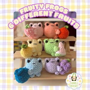 Puede incluir: Exhibición de seis figuras de ranas de ganchillo, cada una con una fruta diferente. Las ranas son de varios colores, incluyendo rosa, naranja, amarillo, verde, azul y morado. El texto "FRUITY FROGS 6 DIFFERENT FRUITS" se muestra encima.