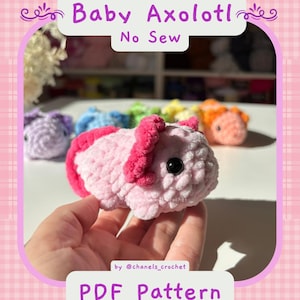 May include: A light pink crocheted baby axolotl plush toy with dark pink gills and black eyes. The image includes text that reads Baby Axolotl No Sew and PDF Pattern by @chanels_crochet. Several other colorful crocheted axolotls are visible in the background.
