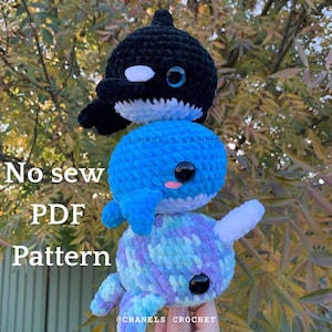 May include: Three crocheted stuffed animals: a black and white orca, a blue whale, and a purple and blue narwhal. The orca is on top of the whale, and the narwhal is on top of the whale. The caption reads "No sew PDF Pattern".