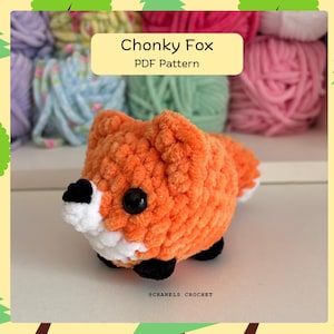 以下が含まれることがあります： オレンジと白の編みぐるみキツネ、黒いアクセント付き。キツネは大きな黒い目を持ち、「Chonky Fox PDF Pattern」と表示されています。背景にはカラフルな糸と木の装飾的なボーダーがあります。