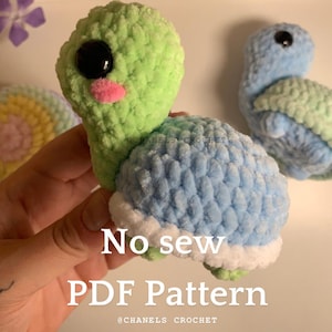 May include: A crocheted turtle amigurumi toy with a green head and a blue shell. The turtle has large black eyes and a pink nose. The toy is made with a no-sew pattern and is available as a PDF download. The text "No sew PDF Pattern" is written on the image.