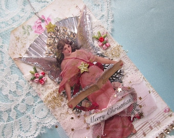 May include: A vintage Christmas ornament featuring a pink angel with gold glitter wings and a star on her dress. The ornament is decorated with lace, glitter, and the words "Merry Christmas".