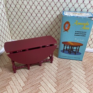 May include: A miniature burgundy drop-leaf table with detailed legs. Beside it is a vintage blue box labeled "Amanda Ann" with text that reads "Exquisite Miniature Furniture". The table is on a wooden floor.