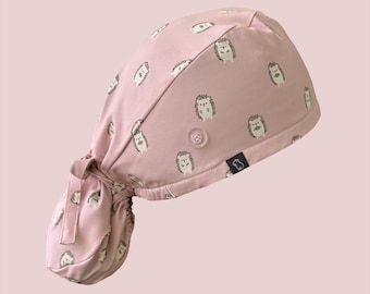 May include: A dusty pink scrub cap featuring a repeating pattern of small, light-colored hedgehogs. The cap includes a side button for securing accessories, an elasticized back, and fabric ties to adjust the fit, with a small black brand tag.
