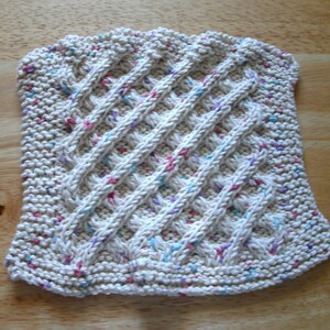 May include: A white knitted dishcloth with a diagonal pattern and speckles of pink, blue, and purple yarn.