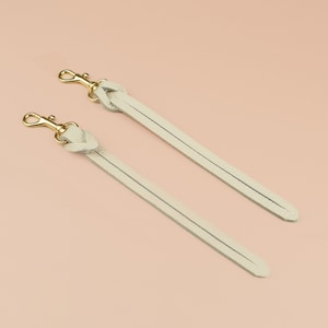 May include: Two cream-colored leather bag straps with gold-tone clasps. Each strap has a knotted detail at the top. The straps are displayed diagonally against a light peach background.
