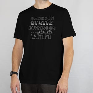 Pode incluir: Camiseta preta com texto branco: "RAISED ON STATIC RUNNING ON WiFi". A camiseta tem gola redonda e mangas curtas. O texto está em uma fonte sem serifa.