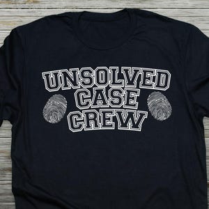 May include: Black t-shirt with the text "UNSOLVED CASE CREW" in white, outlined in black. Two white fingerprint graphics flank the text. The shirt is made of a soft material.