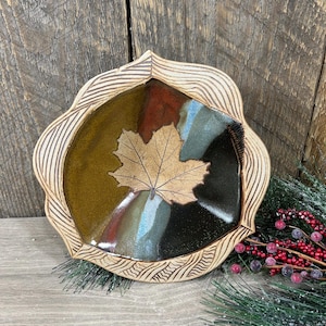 May include: A handcrafted ceramic bowl with a leaf design. The bowl features a maple leaf in the center, surrounded by sections of brown, green, and blue. The rim has a textured, wavy pattern. A decorative piece for the home.