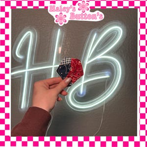 May include: A multi-patterned, geometric-shaped fabric item, held in front of a neon sign that reads "HB". The fabric features a mix of red paisley, blue gingham, and navy patterns. The image is framed with a pink and white checkered border, and includes the text "Haley's Button's".