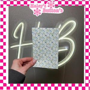 May include: A rectangular piece of paper with a floral pattern of blue and green flowers on a white background. The paper is held in front of a neon sign that says "HB" and a pink and white checkered border.