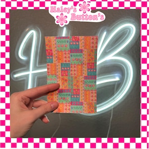 May include: A rectangular piece of patterned fabric with a patchwork design. The fabric features squares of orange, teal, and pink with floral and geometric patterns. The text "Haley's Button's" is visible in the top center.