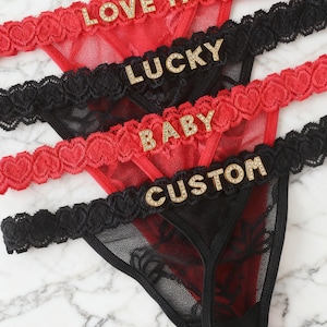 May include: Close-up of three thong underwear in red and black lace. The red thong has the text "LOVE ME" in gold letters. The black thong has the text "LUCKY" in gold letters. The third thong has the text "BABY" and "CUSTOM" in gold letters.