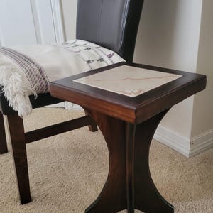 May include: A dark brown, square side table with a light-colored tile top. The table has a unique, curved leg design and is placed next to a black leather chair. A cream-colored throw with a purple pattern rests on the chair.
