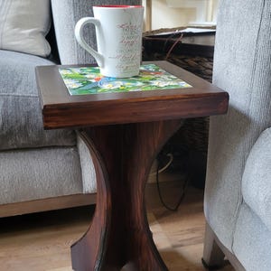 May include: A dark brown wooden side table with a decorative tile top featuring a floral design. A white mug with red trim and text sits on the table. The table has a unique hourglass-shaped base.