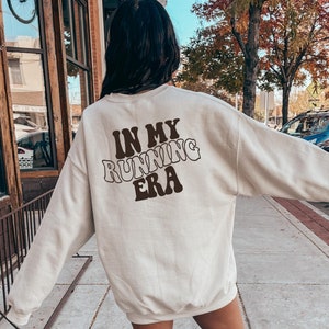 May include: A beige sweatshirt with the text "IN MY RUNNING ERA" in brown letters on the back. The sweatshirt is being worn by a person standing on a sidewalk.
