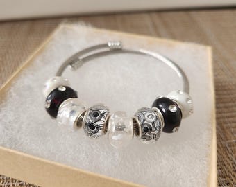 May include: A silver bangle bracelet with decorative beads in black, white, and silver. The beads feature various patterns, including swirls and polka dots. The bracelet is presented in a small, open cardboard box.