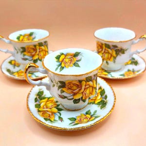 Puede incluir: Tres tazas y platillos blancos con diseños florales de rosas amarillas y ribetes dorados. Las tazas tienen asas curvas y se asientan sobre platillos a juego. El conjunto está dispuesto sobre una superficie rosa claro.