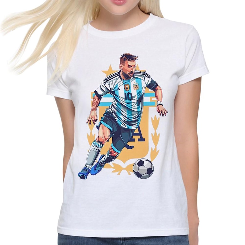 May include: White t-shirt with a graphic of a soccer player in a blue and white jersey with the number 10 on the front. The player is dribbling a soccer ball and is surrounded by a gold crest with the letters "AFA".