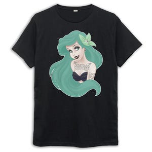 Puede incluir: Camiseta negra con una ilustraci&oacute;n de una sirena de pelo verde. La sirena tiene tatuajes y una flor en el pelo. El texto "Lost at Sea" es visible en su brazo.