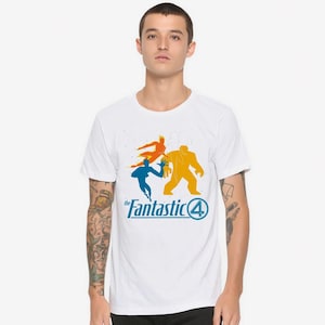 May include: White t-shirt with a graphic of the Fantastic Four. The design features orange, blue, and yellow character silhouettes, and the words "the Fantastic 4" in blue. The shirt is made of a soft material.