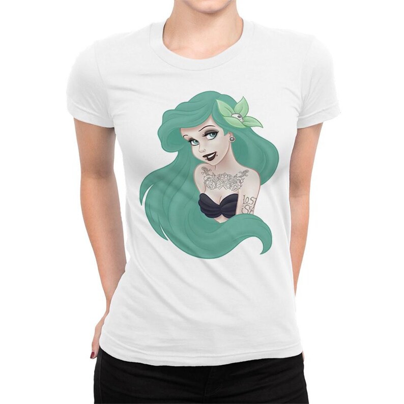Puede incluir: Una camiseta blanca con un gr&aacute;fico de una sirena de pelo verde con tatuajes y un top de bikini negro. La sirena tiene una flor en el pelo y un pendiente de calavera.
